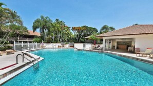 A clear blue pool with a white deck, surrounded by palm trees and a building with a tiled roof.