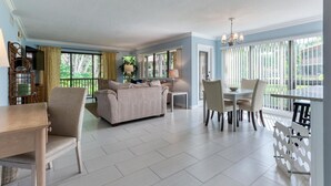 A spacious living room with a beige sofa, dining table, and chairs. There are large windows with curtains, a chandelier, and a view of greenery outside.