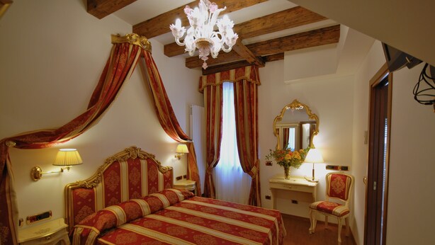 Een slaapkamer met een groot bed, een nachtkastje met lamp, een stoel, een kaptafel met spiegel en een kroonluchter.