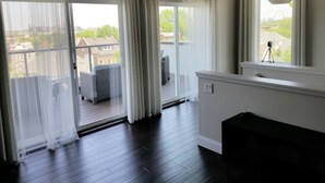 Ein Zimmer mit Balkon, großen Fenstern mit weißen Vorhängen, Holzboden und einem kleinen Tisch.