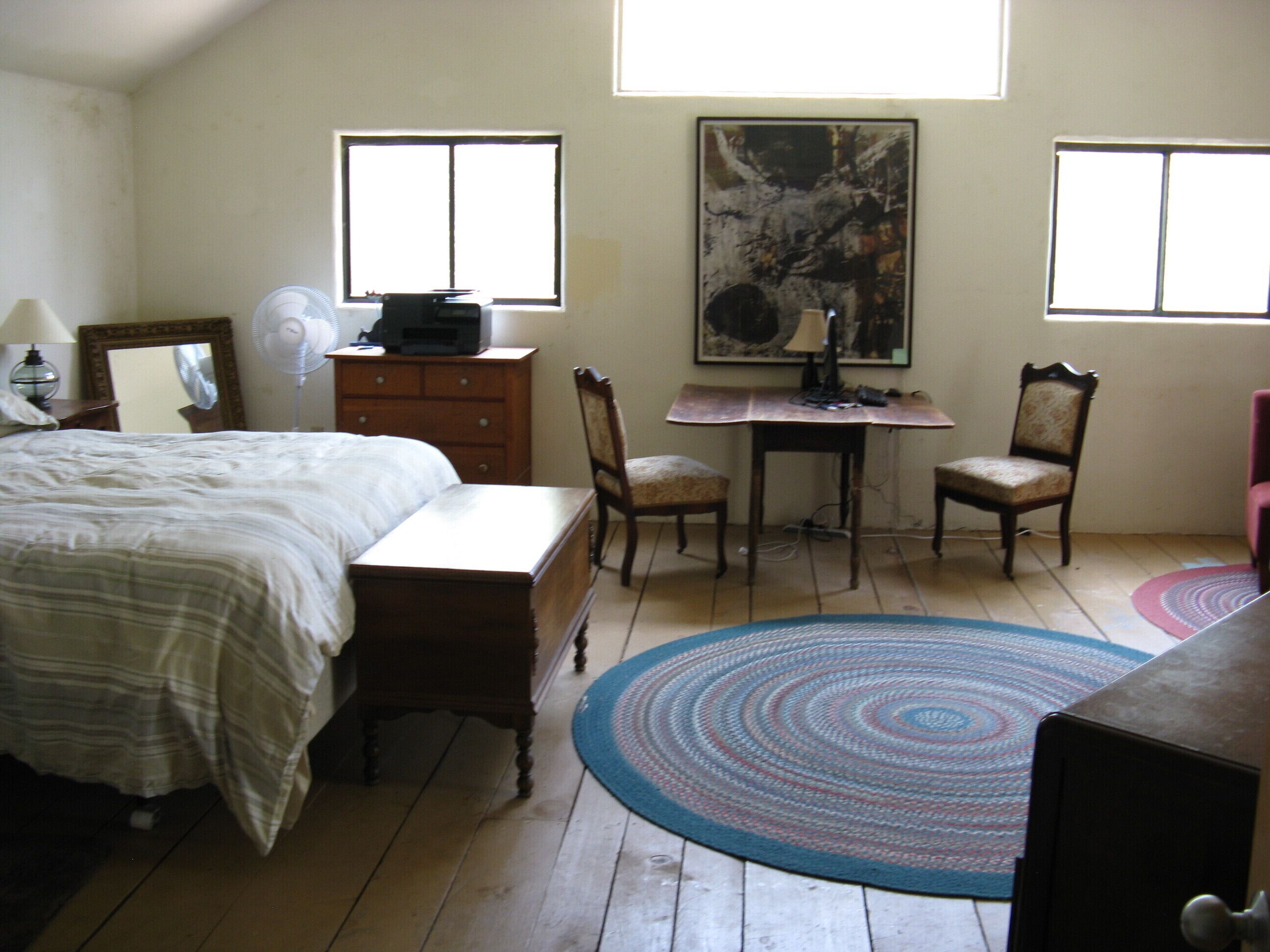 Un dormitorio con cama, cómoda, escritorio y sillas. Hay un cuadro en la pared y una ventana que proporciona luz natural.
