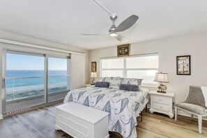 Une chambre avec une grande fenêtre, un ventilateur de plafond, un lit avec une literie à motifs, une table de chevet avec une lampe et une chaise.