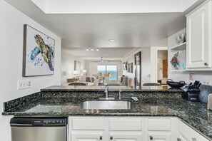 A modern kitchen with a granite countertop, stainless steel sink, and white cabinetry. There is a dishwasher and a dining area visible in the background. - 🏖️Escape to this Gulf front 2 BR / 2 BTH condo with swimming pool (Pensacola Beach)