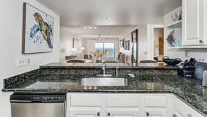 A modern kitchen with a granite countertop, stainless steel sink, and white cabinetry. There is a dishwasher and a dining area visible in the background. - 🏖️Escape to this Gulf front 2 BR / 2 BTH condo with swimming pool (Pensacola Beach)