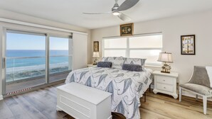 A bedroom with a large window, a ceiling fan, a bed with patterned bedding, a bedside table with a lamp, and a chair. - 🏖️Escape to this Gulf front 2 BR / 2 BTH condo with swimming pool (Pensacola Beach)