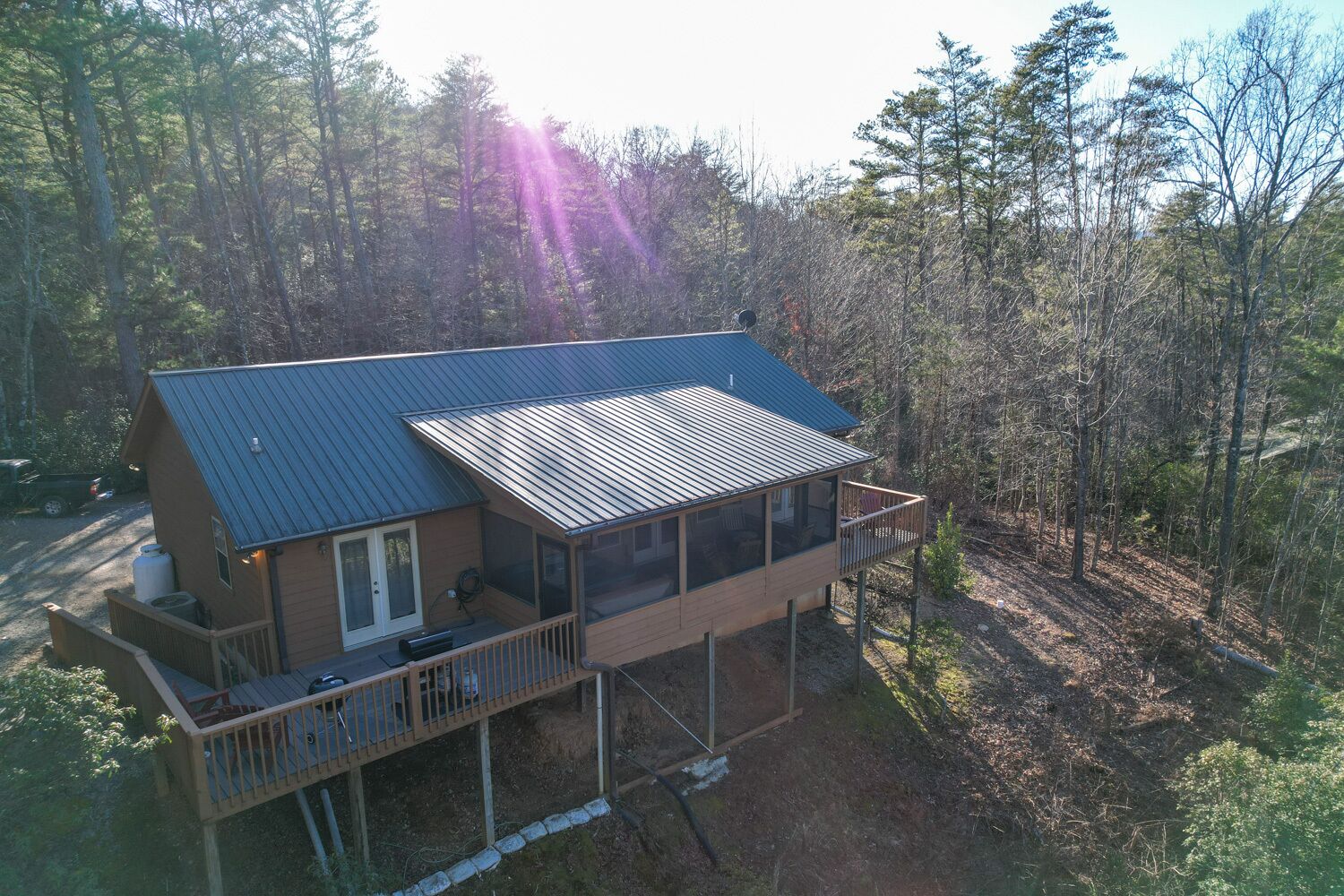 A two-story cabin with a metal roof, a deck, and a large window overlooking a forested area.