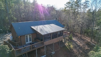 A two-story cabin with a metal roof, a deck, and a large window overlooking a forested area.