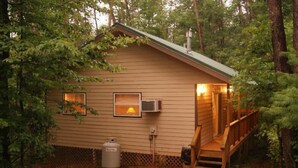 A small cabin with a wooden deck, a chimney, and a window with a light on.