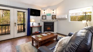 A living room with a fireplace, a television mounted on the wall, and a large window with a view of trees.