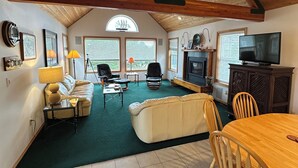 Een ruime woonkamer met een hoog houten plafond, een open haard, een televisie en een eettafel met stoelen.