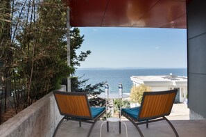 Ein Balkon mit zwei Holzstühlen und einem kleinen Tisch, der Blick auf die Küste mit blauem Himmel und fernen Bergen bietet.