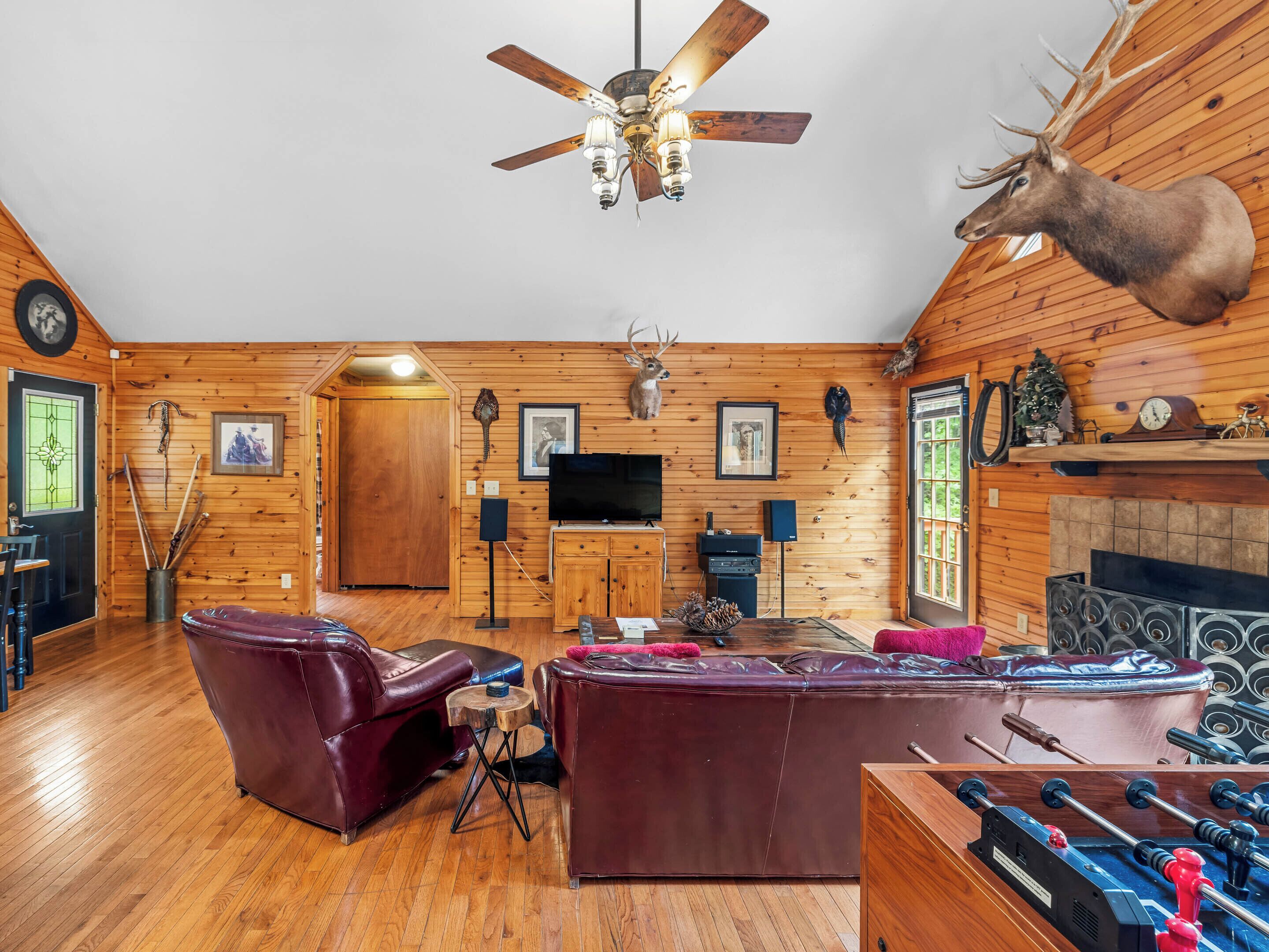 A rustic cabin interior with wooden walls, a ceiling fan, a mounted deer head, a fireplace, leather sofas, and a foosball table.