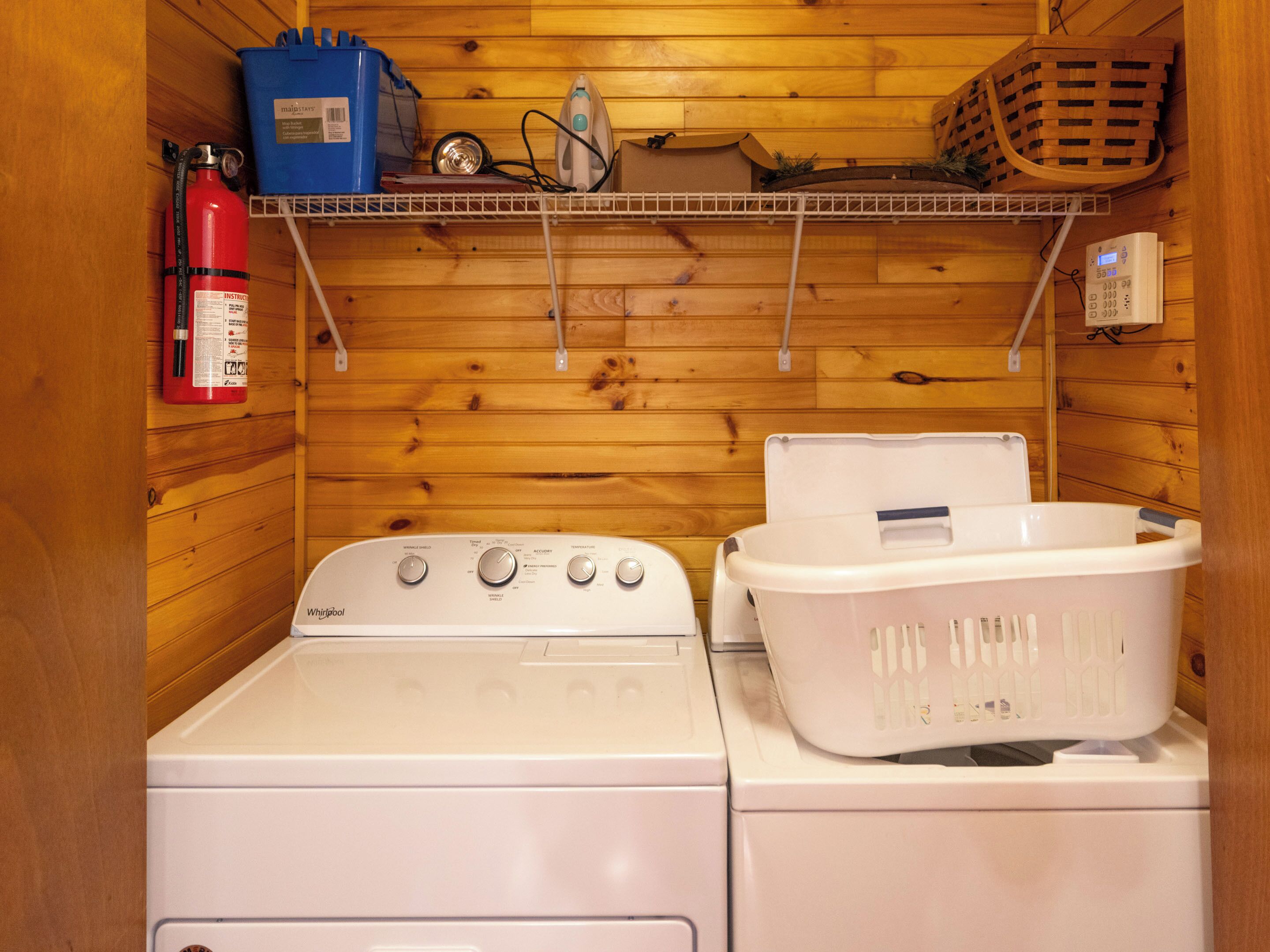 A laundry room with a Whirlpool washer and dryer, a fire extinguisher, a blue storage bin, and a basket on a shelf.