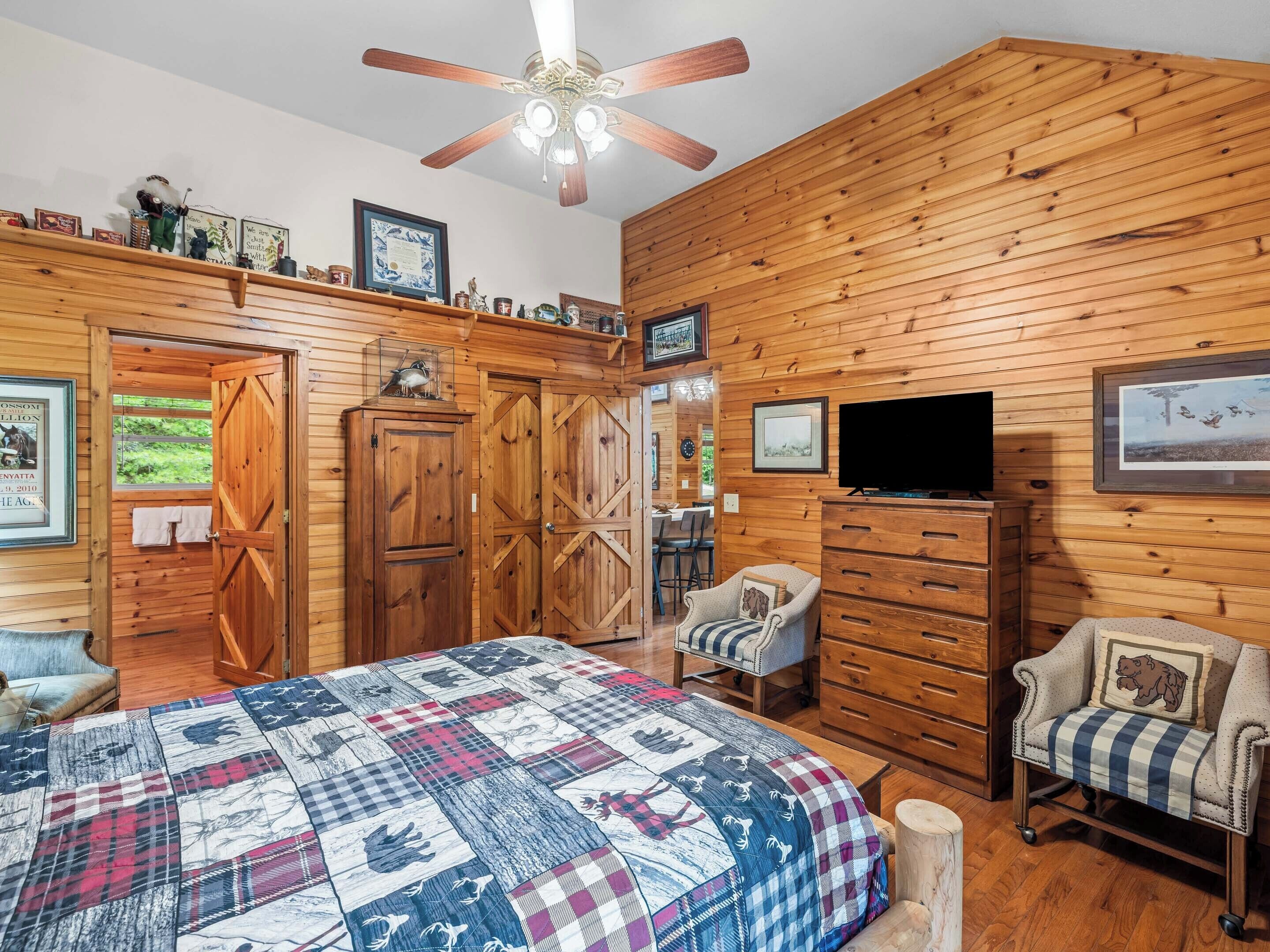 A wooden-paneled room with a bed, two chairs, a TV, and a ceiling fan.