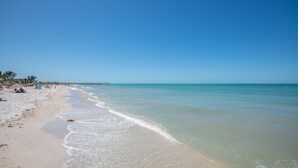 Une plage avec une eau d’un bleu limpide, un rivage sablonneux et quelques personnes visibles au loin.