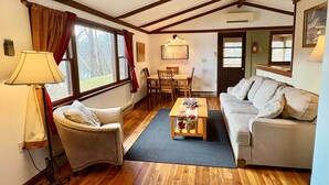 A cozy living room with a sofa, armchair, dining table, and a view of the outdoors through large windows. - Stepping Stone Cabin (Page County)