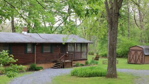 A rustic cabin with a porch, a small deck, and a shed in a wooded area. - Stepping Stone Cabin (Page County)