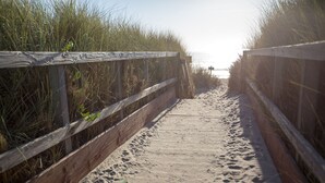 Ein hölzerner Steg führt zu einem Strand, im Sand sind Fußabdrücke zu sehen.