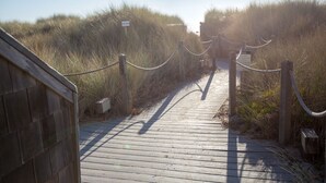Ein hölzerner Steg führt zu einem Strandbereich mit einem Seilabfangnetz und einem Schild, das Strandregeln aufzeigt.