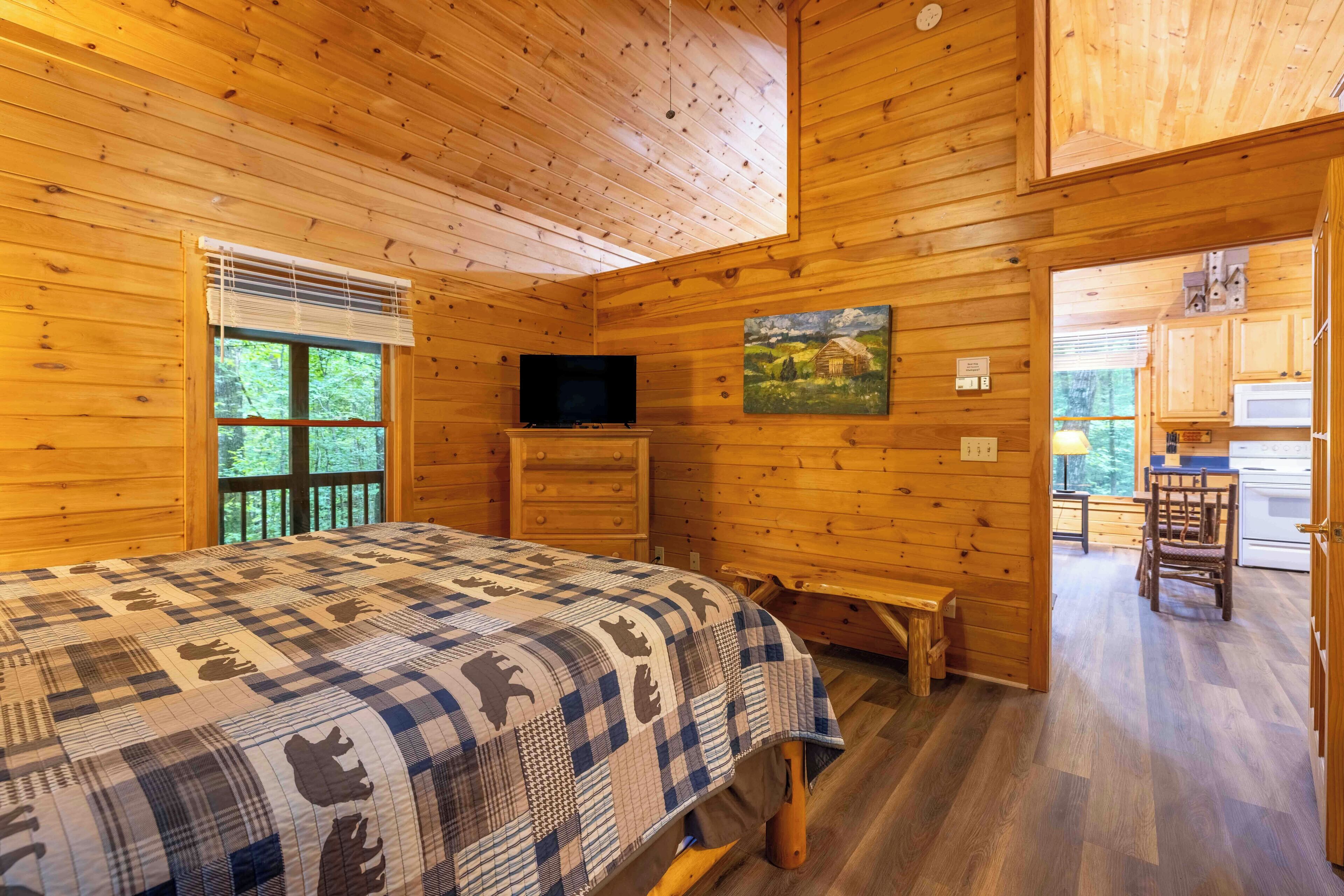 A wooden cabin room with a bed, a television, a painting, and a kitchen area visible through an open door.
