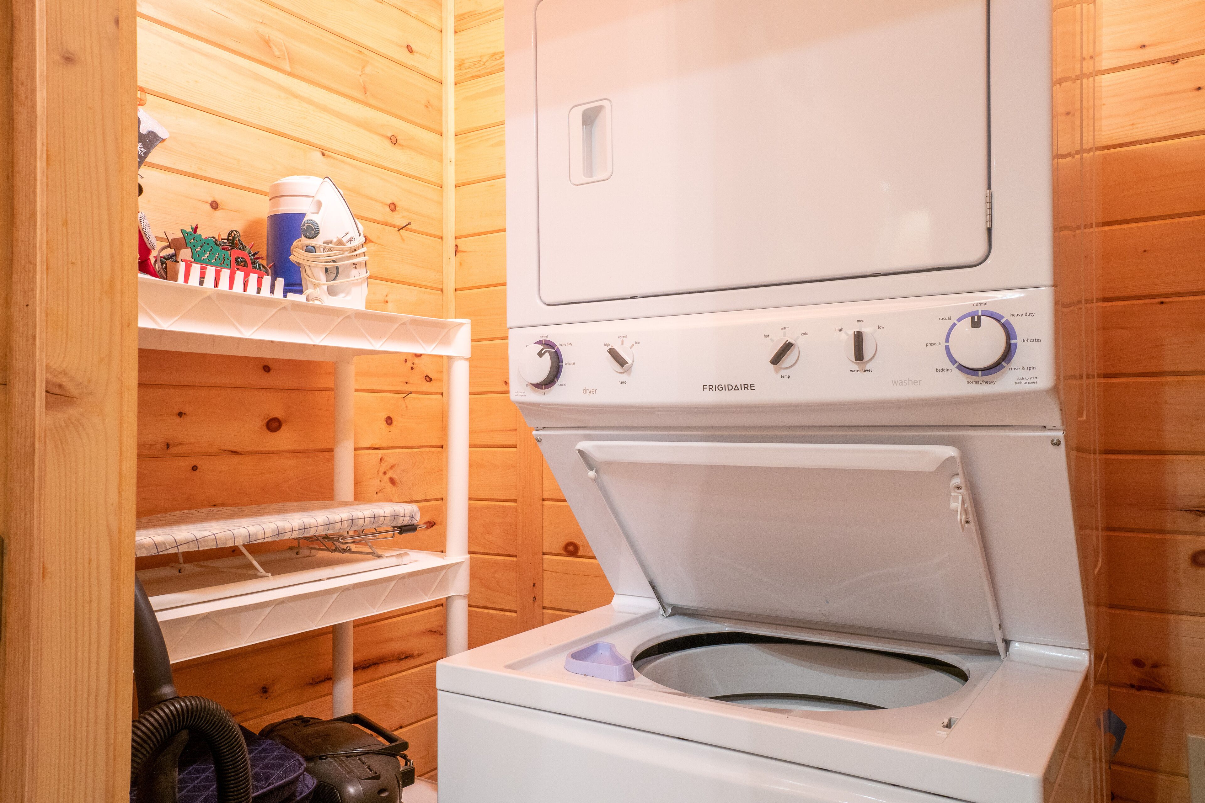 A compact, white, two-door washer and dryer unit in a wooden cabin with a shelf above containing various items.
