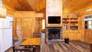 A wooden cabin interior with a stone fireplace, wooden cabinets, a dining table, and a television mounted above the fireplace. - Bear Hug - Endorsed by Theodore Bear, a.k.a. "Teddy" (Sautee Nacoochee)