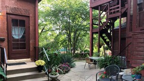 A brick building with a wooden staircase leading to a door, a patio area with outdoor furniture, and a staircase on the right side. - Vineyard Mansion - Carriage House - 1 Queen 1 Full Bed (St Joseph)