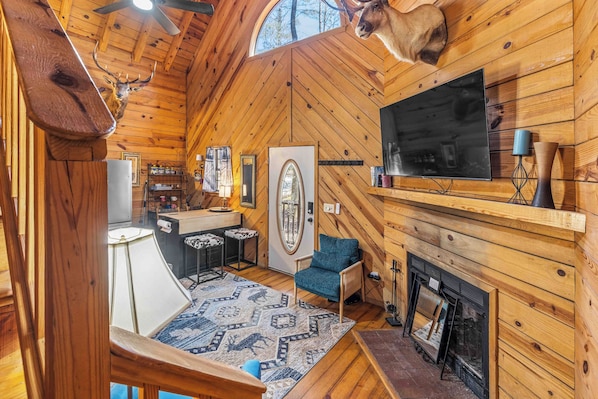 Interior de cabana de madeira com cabeça de veado montada na parede, lareira, televisão e área de estar aconchegante.