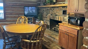A rustic wooden cabin interior with a round wooden dining table surrounded by chairs, a stone fireplace, wooden cabinets, and a television mounted on the wall. - Lake Barkley Log Cabin Getaway + Free Weekday Pontoon (seasonal)  (Cadiz)