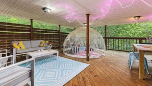Un espacio exterior techado con terraza de madera, un sofá blanco con cojines de colores, una mesa, sillas y una estructura de domo geodésico.