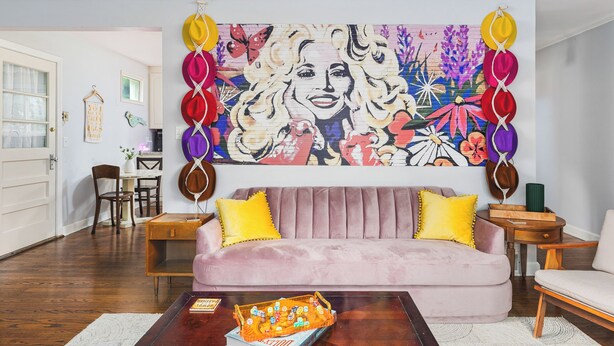 Un salón con un mural grande y colorido, un sofá rosa, almohadas amarillas y una mesa de centro con una revista.