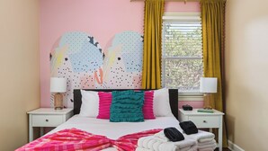 Un dormitorio con cama, almohadas, una mesita de noche, una lámpara, una ventana con cortinas y un mural en la pared.