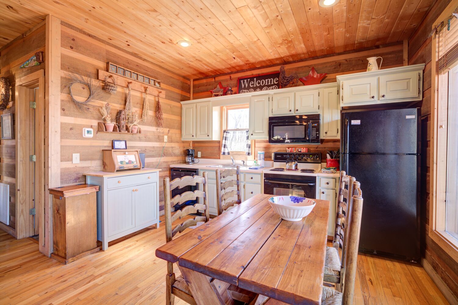 A rustic kitchen with wooden walls, a wooden ceiling, and a central island with a bowl. There are white cabinets, a black refrigerator, and a microwave above the island.