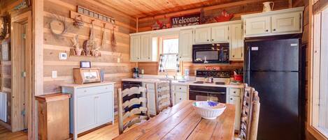 A rustic kitchen with wooden walls, a wooden ceiling, and a central island with a bowl. There are white cabinets, a black refrigerator, and a microwave above the island.
