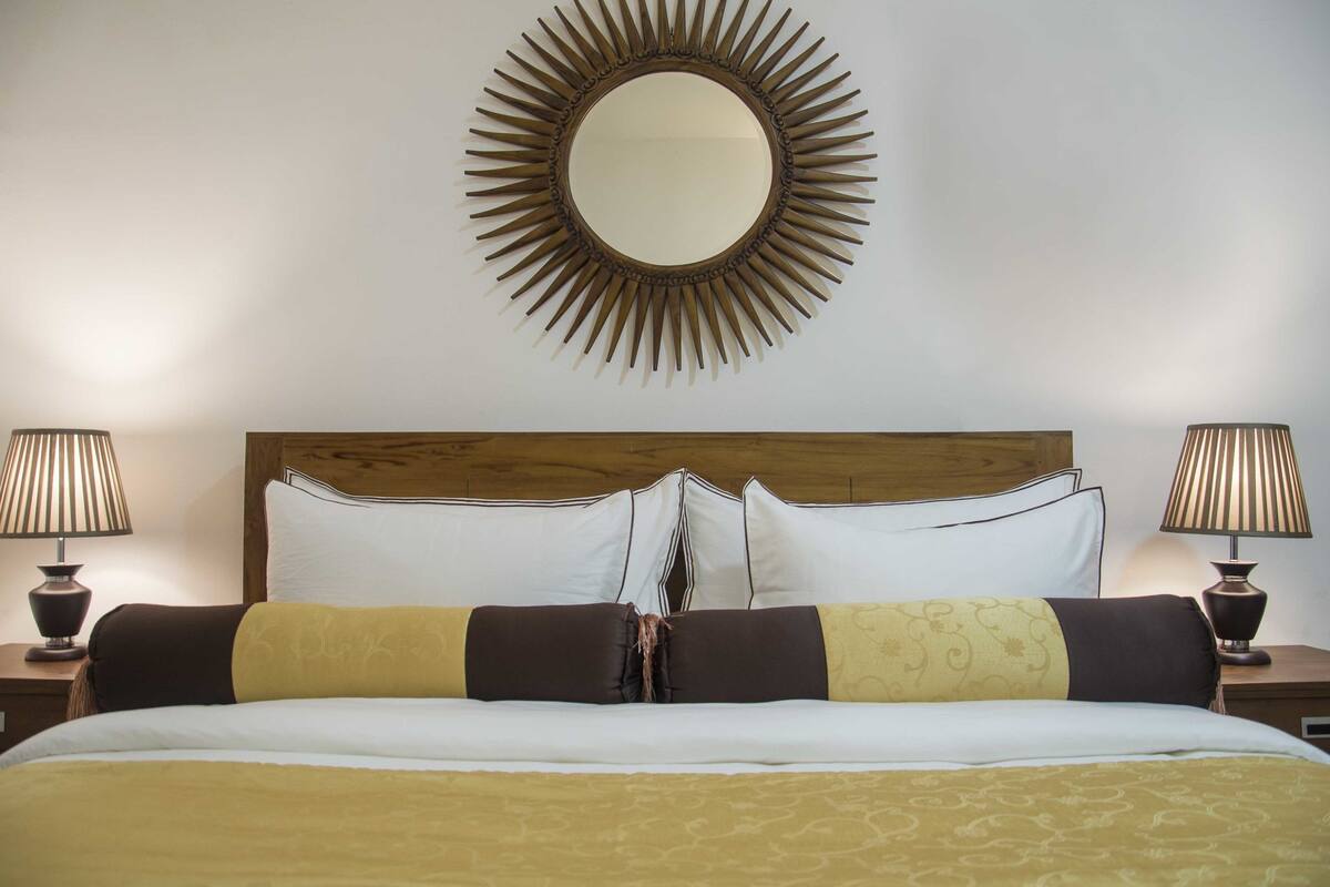 Quarto de hotel com uma cama grande, duas luminárias de cabeceira, uma cabeceira de madeira e um espelho em formato de sol na parede.