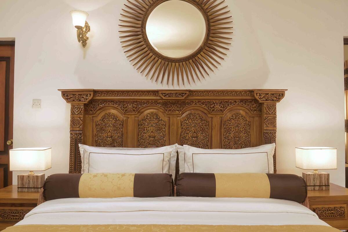 Quarto de hotel com uma cama grande, cabeceira ornamentada, duas luminárias de apoio e espelho fixado na parede.