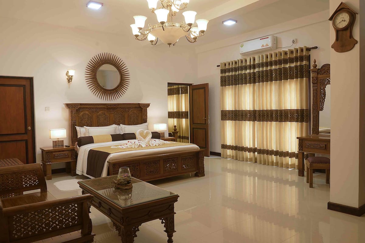 Quarto de hotel com uma cama grande, um candelabro, um espelho, uma cômoda de madeira e uma mesa de cabeceira com abajur.