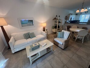 A living room with a white sofa, a coffee table, wicker chairs, a dining table, and a kitchen in the background.
