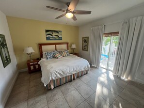 A bedroom with a bed, a nightstand, a ceiling fan, a window with curtains, and a painting on the wall.