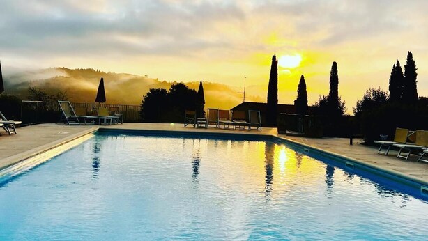 Una piscina con lettini e ombrelloni, circondata da alberi e un tramonto sullo sfondo.