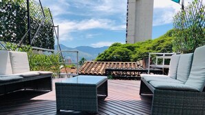 A wooden deck with wicker furniture, a glass table, and a view of mountains and a building in the background.