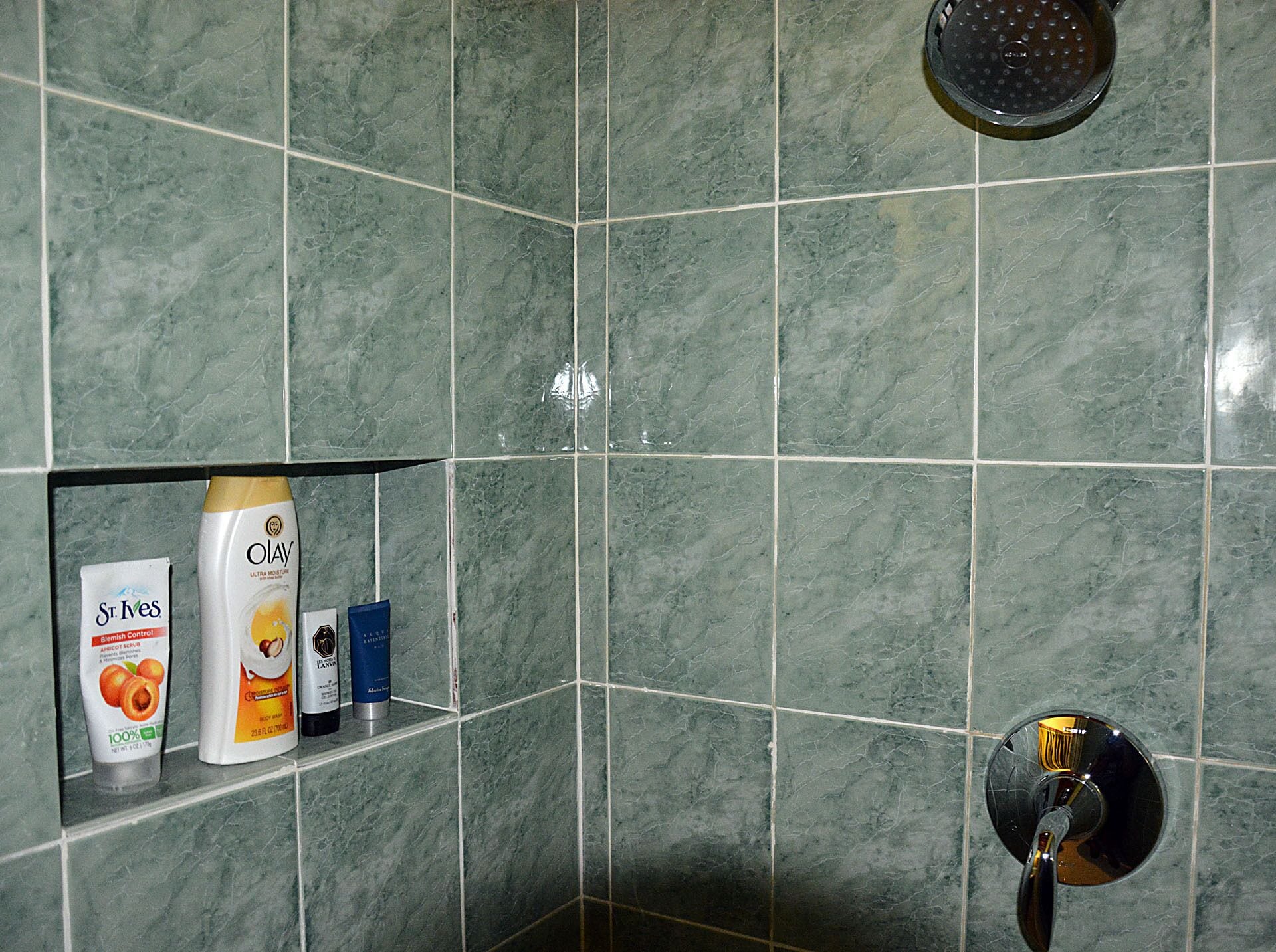 Une salle de bain avec des carreaux de marbre vert, une pomme de douche et des articles de toilette, notamment un flacon de produit Olay, une boîte de produits St. Ives et un flacon bleu.