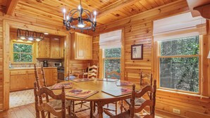 Interno di una baita in legno con un tavolo da pranzo, sedie, un lampadario e un angolo cottura.