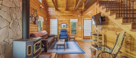 Een gezellig interieur van een chalet met een stenen muur, houten vloeren, een open haard, een bank, een fauteuil, een televisie en een trap.