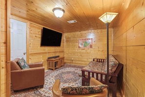 A wooden interior with a flat-screen TV mounted on the wall, a sofa, a patterned rug, and a wooden floor lamp.