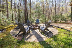Ein kreisförmiger Sitzbereich im Freien mit Adirondack-Stühlen um ein Feuerbecken, umgeben von bewaldeter Landschaft.