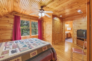 A wooden cabin bedroom with a bed, a ceiling fan, a television, and a view of the outdoors through a window.