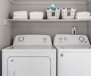 A laundry area with two washing machines and a shelf above containing folded towels and laundry baskets. - Located In The Heart Of OCC with Spectacular Mountain Views in Every Direction! (Colorado Springs)