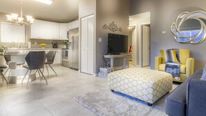 A modern apartment with a dining area, kitchenette, living room with a sofa and ottoman, and a TV mounted on the wall.