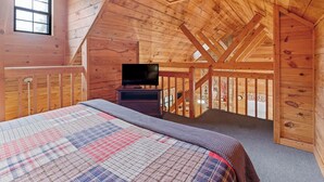 A cozy attic bedroom with a wooden ceiling, a bed with a plaid comforter, a television on a stand, and a window providing natural light. - Country Charm A - Pet Friendly Group Cabin (Helen)